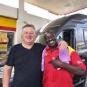 Zwei lächelnde Männer, einer in Shell-Uniform, vor einem Van an einer Tankstelle.
