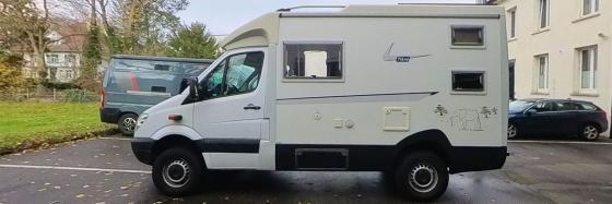 Ein weißer Wohnmobil-Transporter steht auf einem asphaltierten Parkplatz vor einem Gebäude, umgeben von Bäumen.